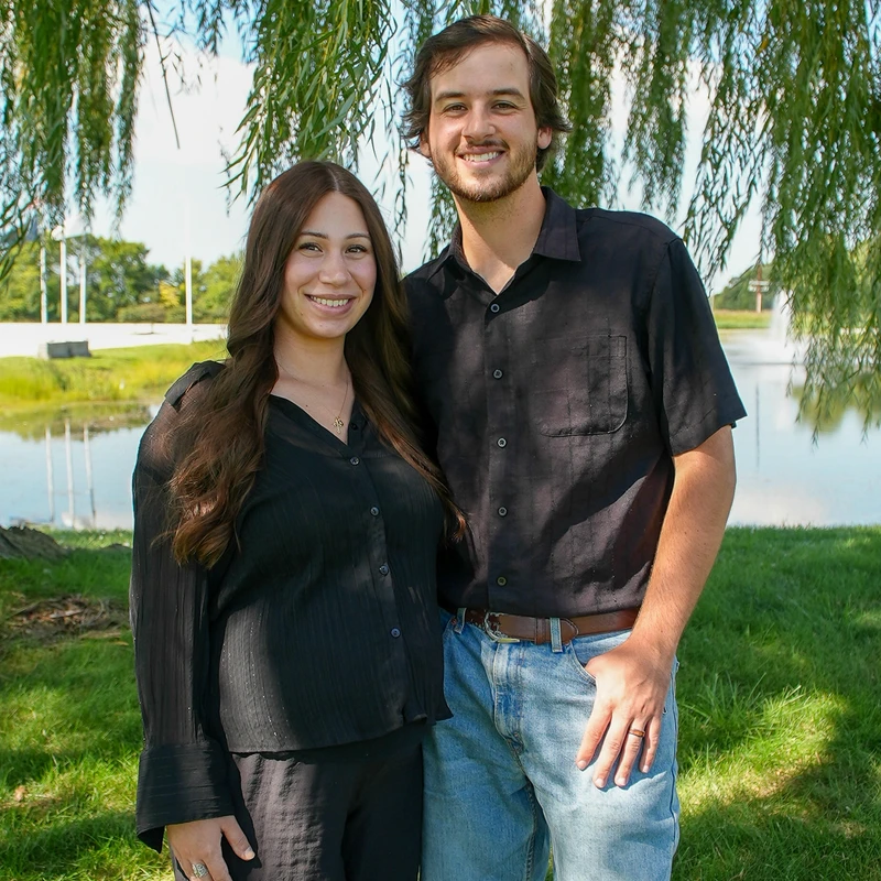 A smiling young couple stands close together under the drooping branches of a willow tree. They are outdoors in a grassy park with a pond in the background.